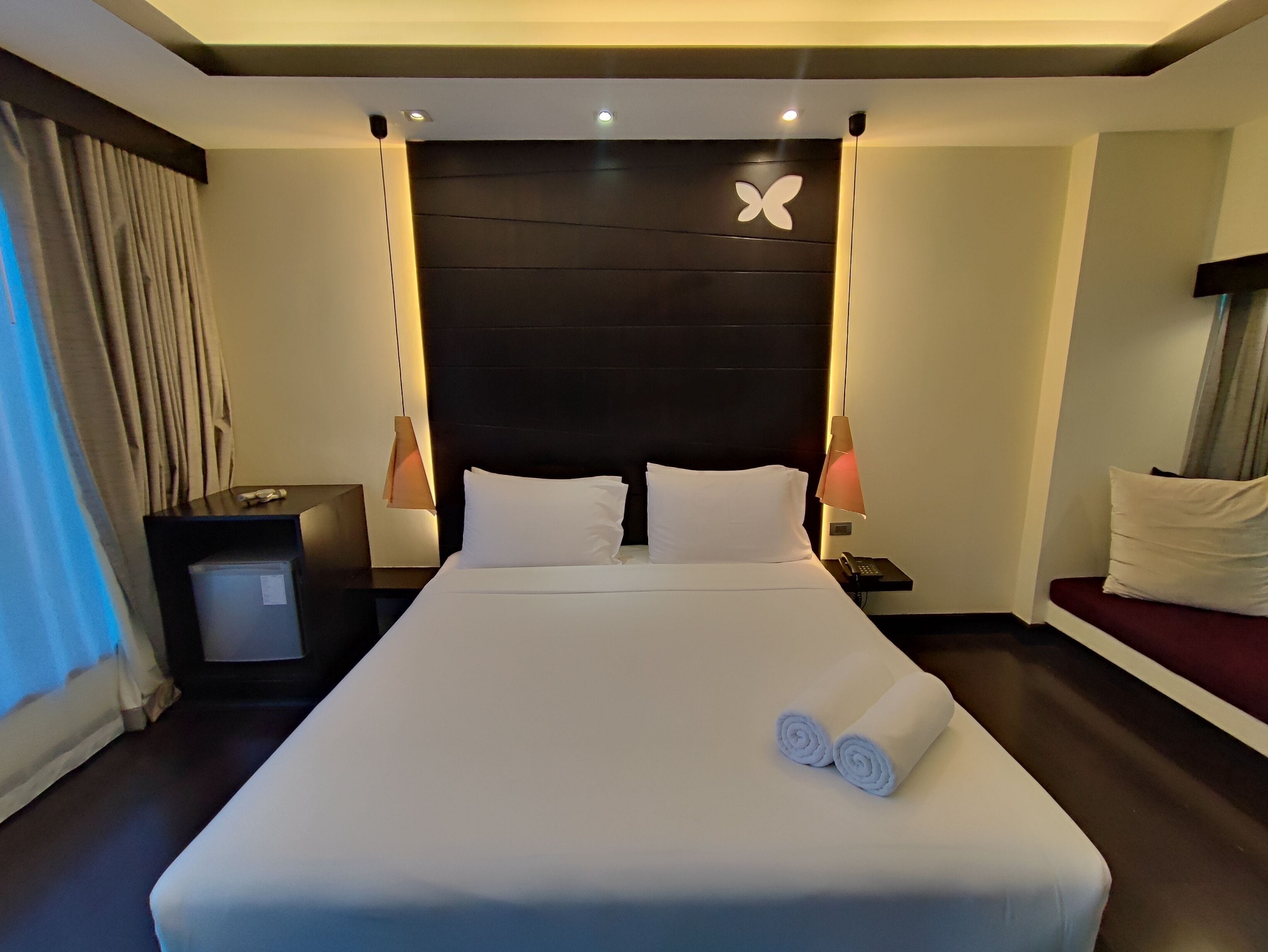 A hotel room with a large bed, two bedside tables, a nightstand with a lamp, a wall-mounted telephone, and a window with curtains.