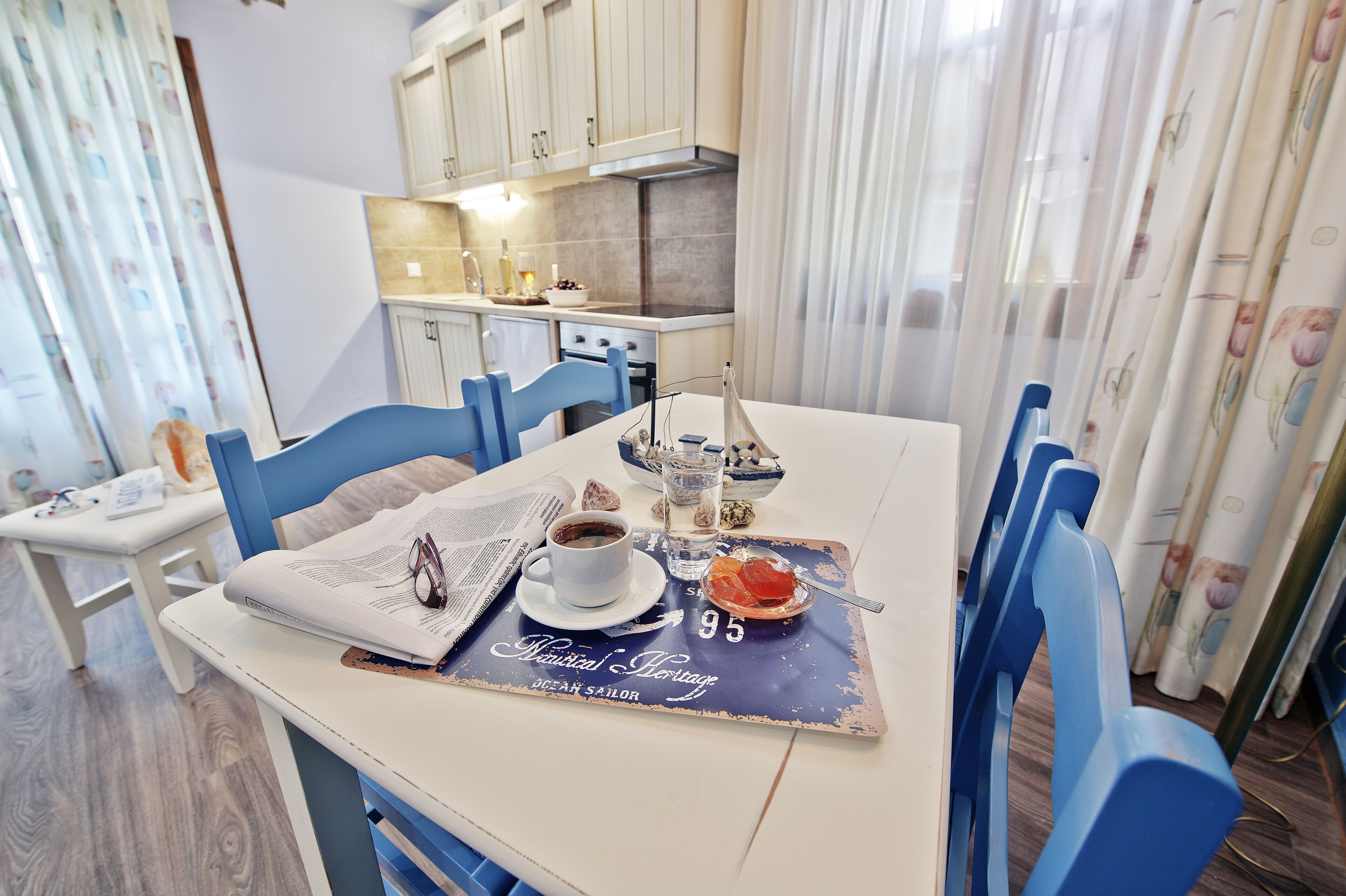 A dining area with a table set for two, a coffee cup, a bowl of fruit, and a menu. The room has light-colored walls, a kitchen in the background, and blue chairs.