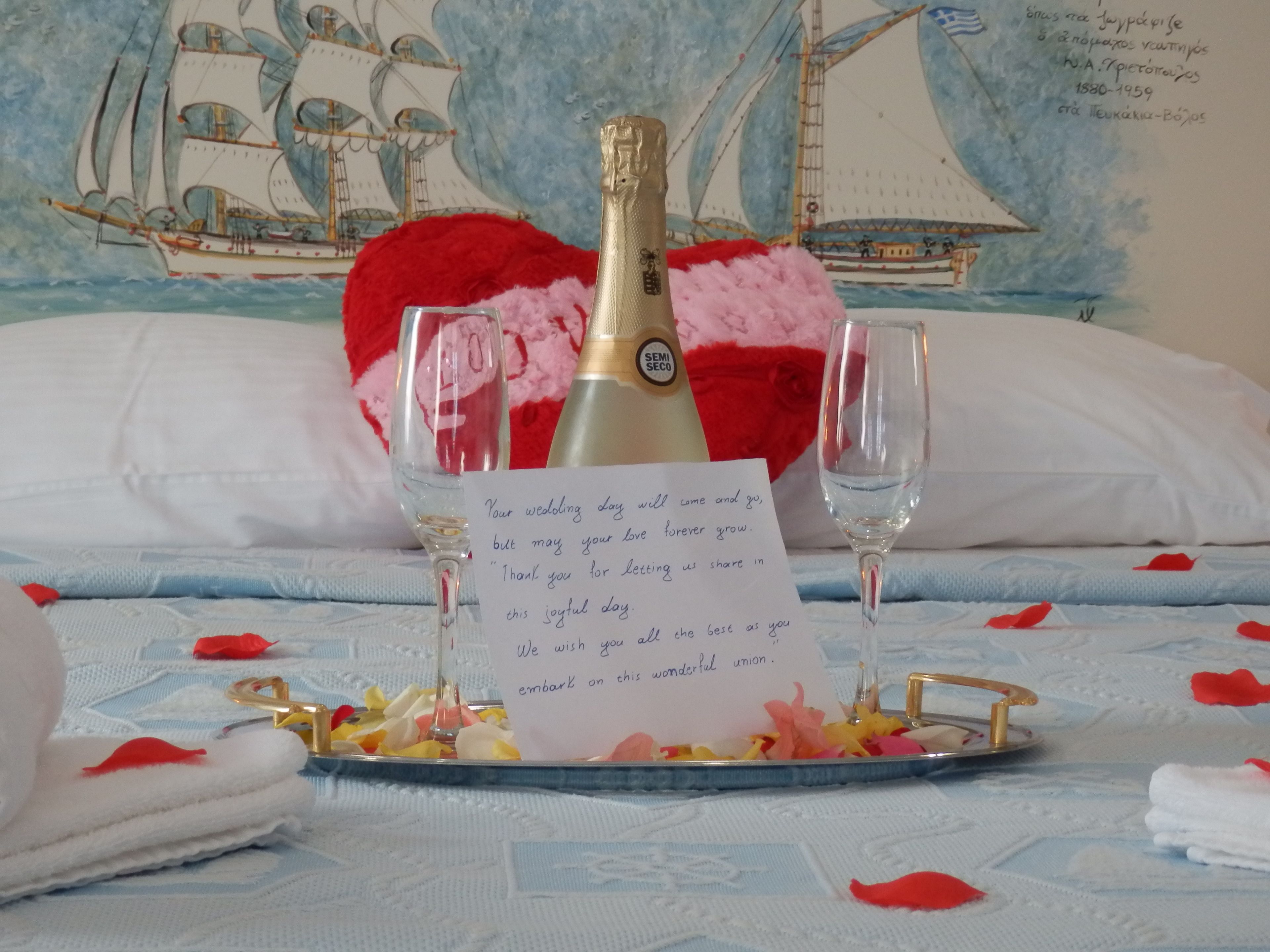 A bed with a decorative sailboat painting, a red heart pillow, a bottle of champagne, two wine glasses, and a note on a tray with rose petals.