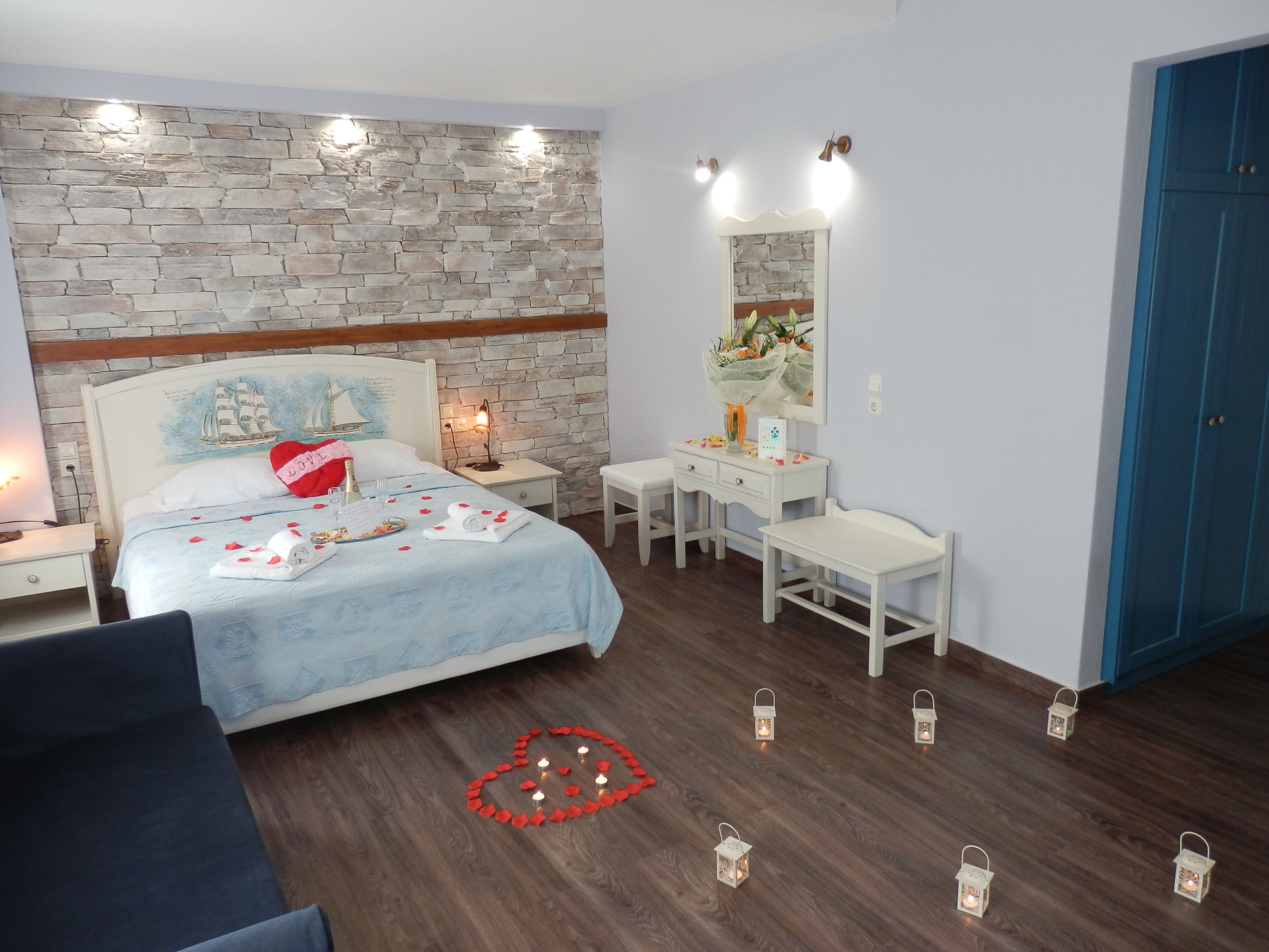 Une chambre avec un mur en pierre, un lit agrémenté d’un oreiller en forme de cœur, des tables de chevet, une commode, un miroir et une porte bleue.