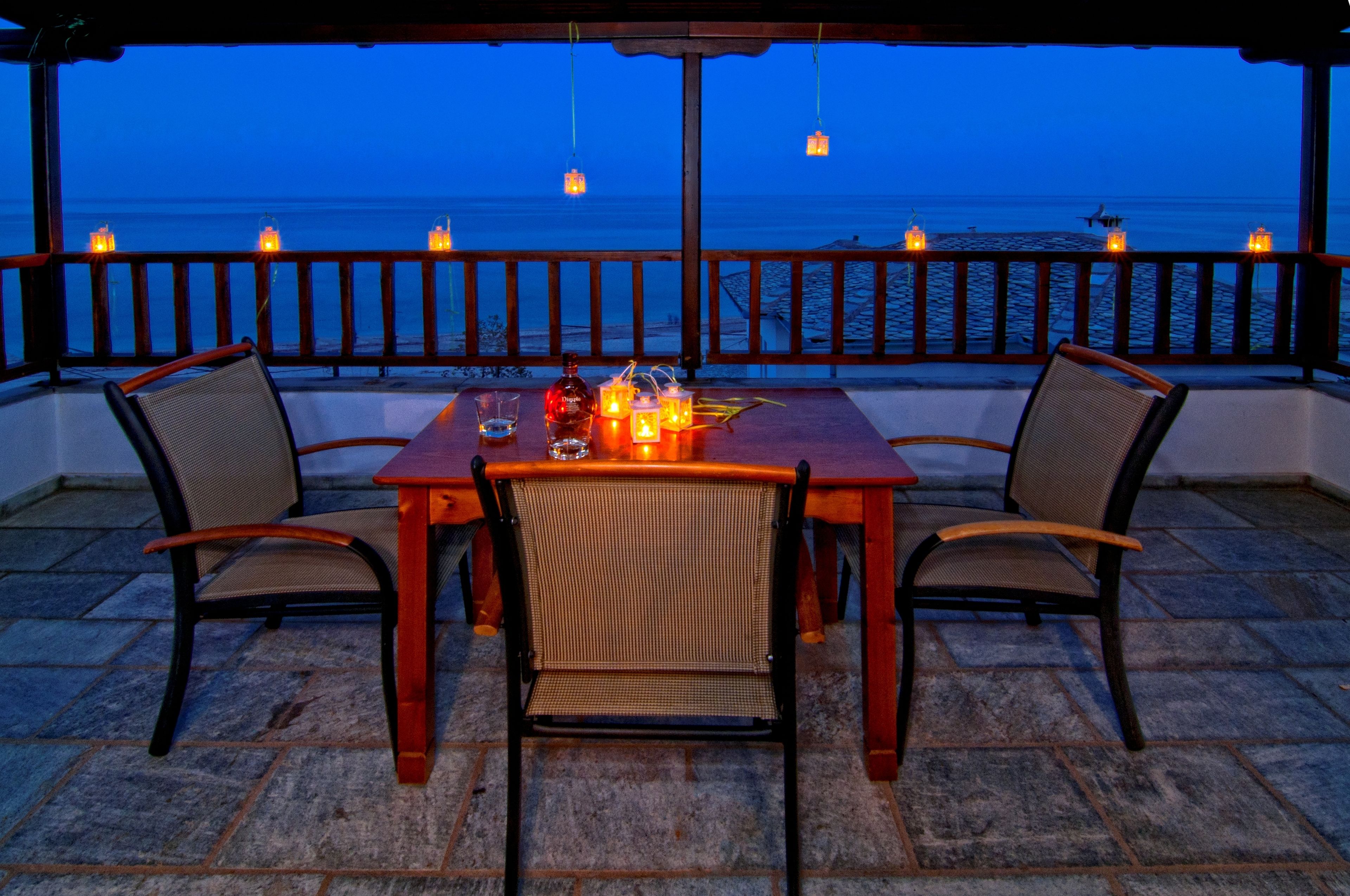 Een houten tafel met twee stoelen, kaarsen en een fles op een balkon met uitzicht op zee.