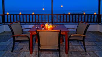 Een houten tafel met twee stoelen, kaarsen en een fles op een balkon met uitzicht op zee.