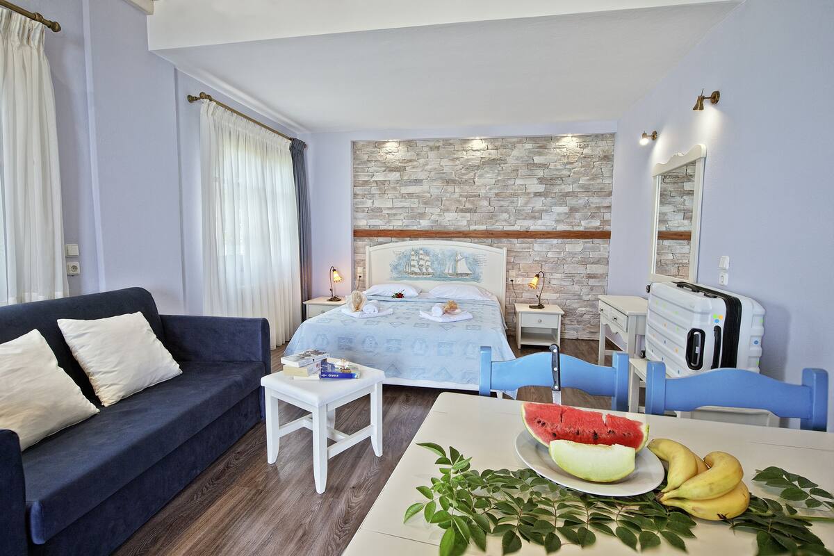 Un dormitorio con cama, mesita de noche, cómoda, silla, sofá y mesa de comedor con frutas y una sandía.