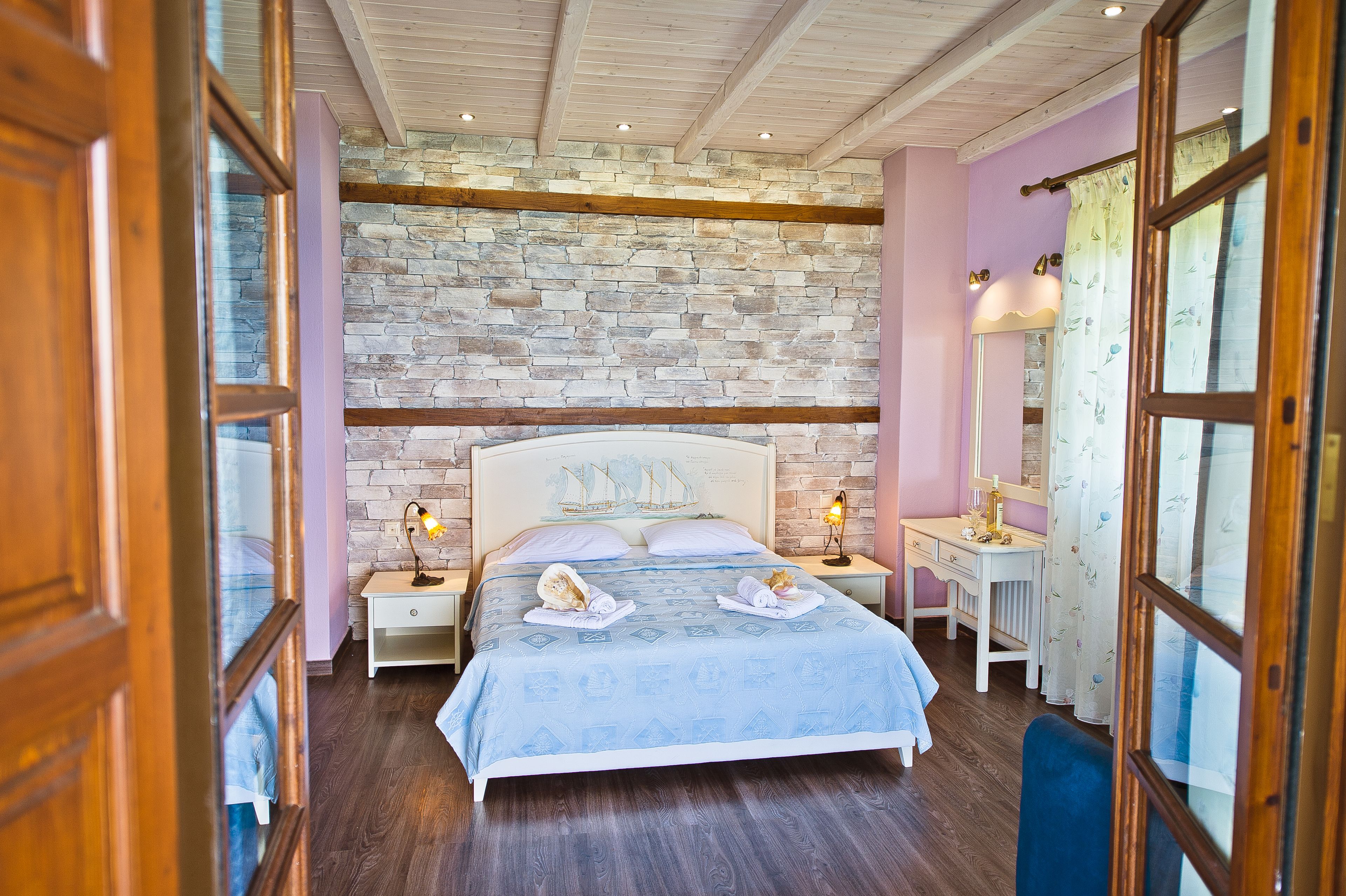 Un dormitorio con pared de piedra, vigas de madera en el techo, una cama con colcha azul, una mesita de noche con lámpara y un escritorio con silla.