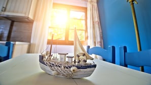 Une maquette de bateau est posée sur une table dans une pièce aux murs bleus, avec une fenêtre ornée de rideaux.