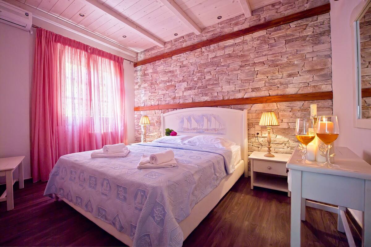 Un dormitorio con pared de piedra, una cama con colcha azul estampada, una mesita de noche con dos lámparas y una pequeña mesa con dos copas de vino.