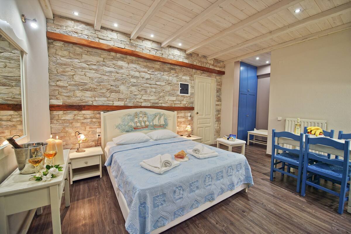 Un dormitorio con pared de piedra, vigas de madera, cama con toallas, mesita de noche, mesa con lámpara y una silla.