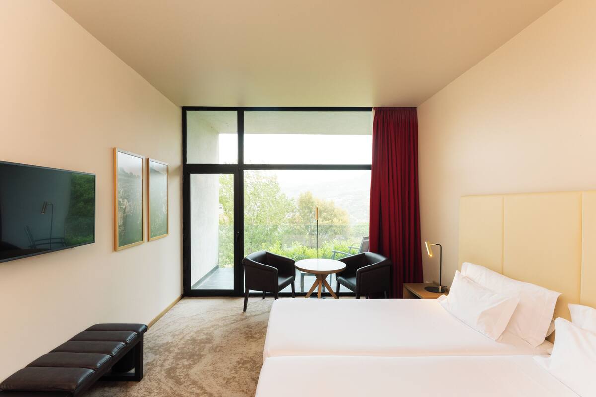 A hotel room with a large bed, a TV on the wall, a seating area with chairs, and a view through the window.