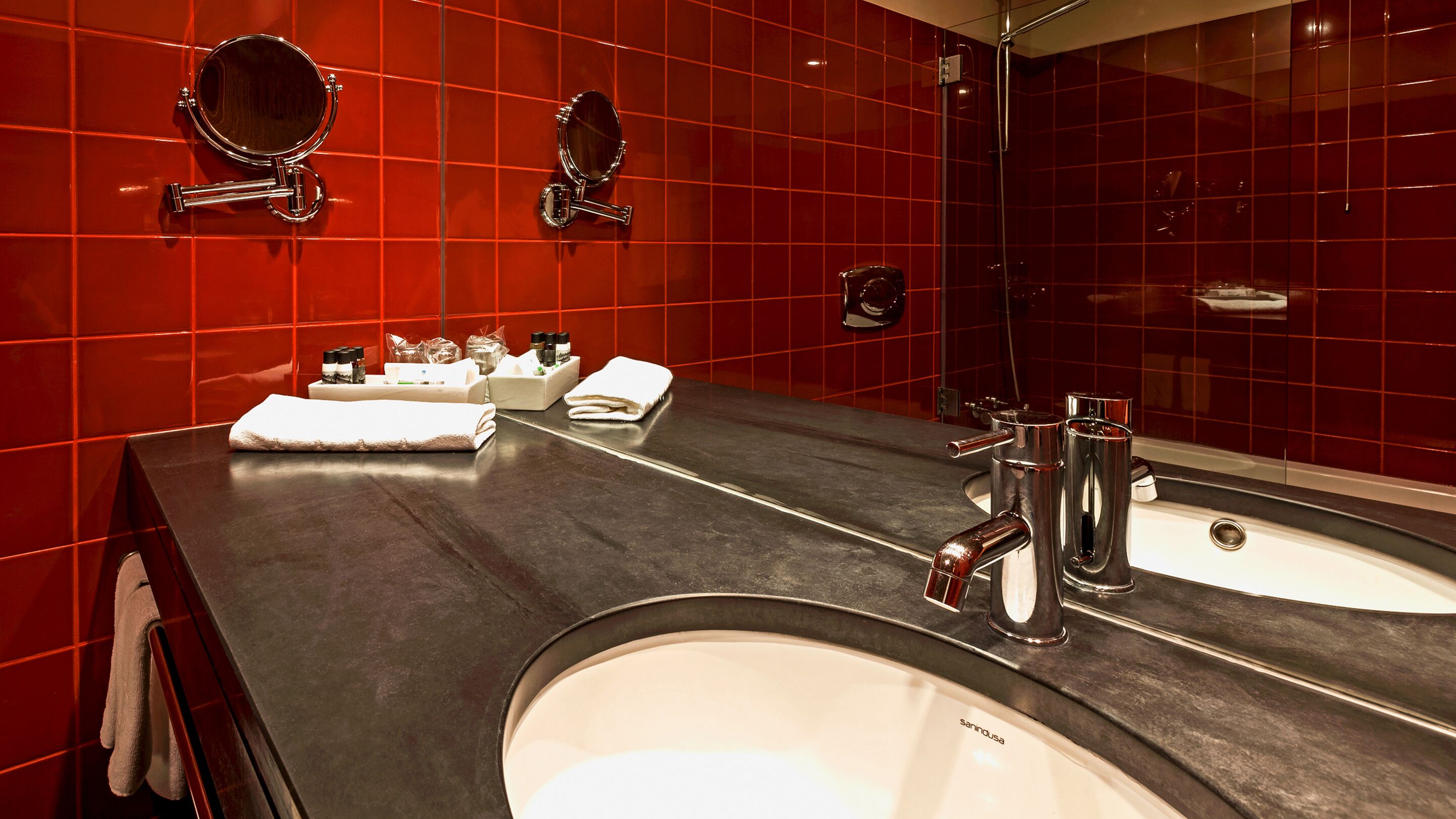 Un baño moderno con azulejos rojos, un espejo grande, dos lavabos y artículos de aseo.