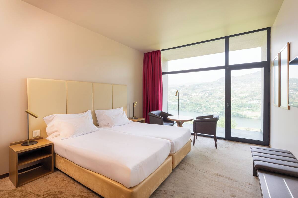 A hotel room with two beds, a desk, a chair, and a view of the outdoors.