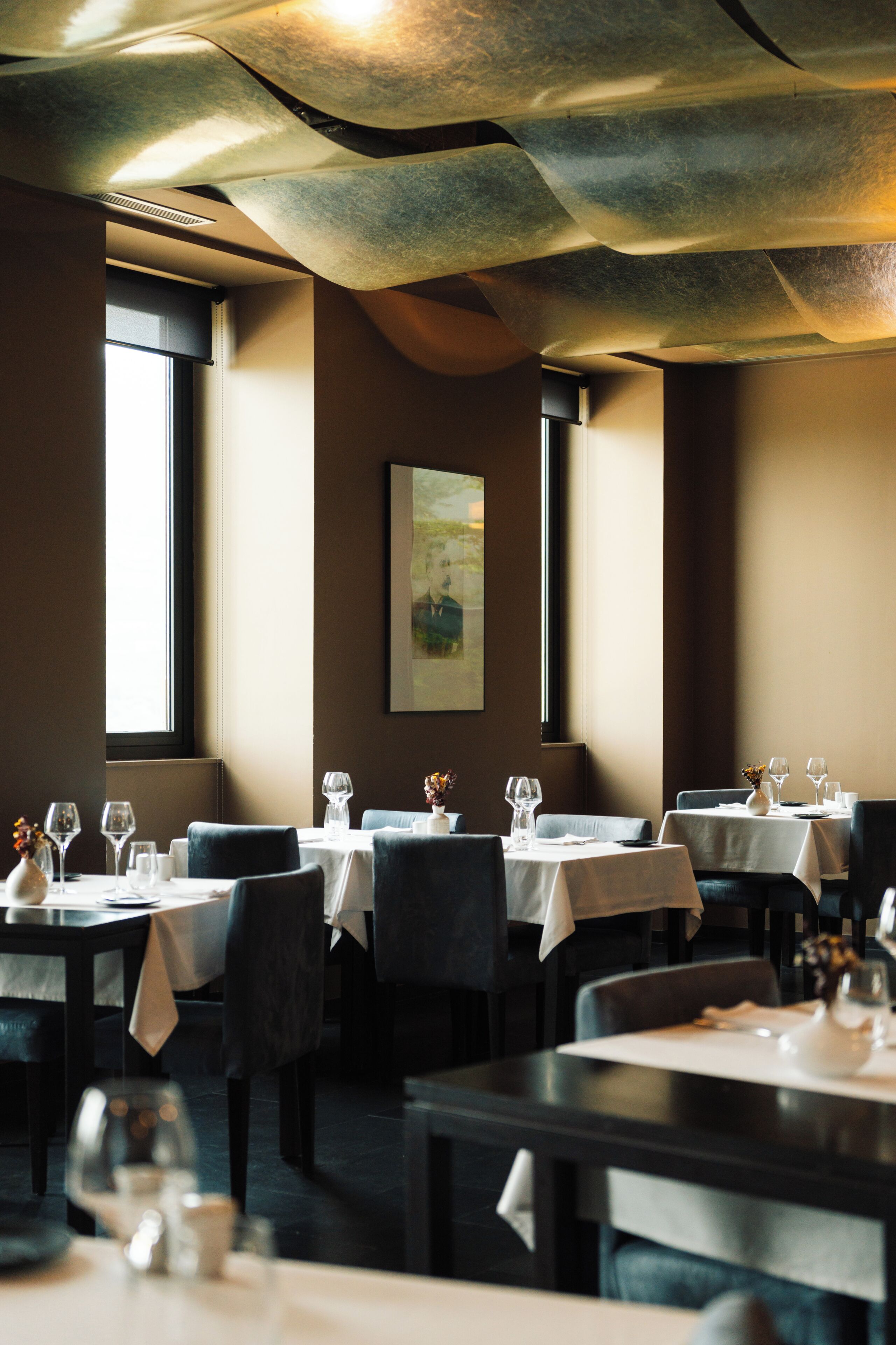 Een restaurant met gedekte tafels, een schilderij aan de muur en grote ramen voor natuurlijk licht.