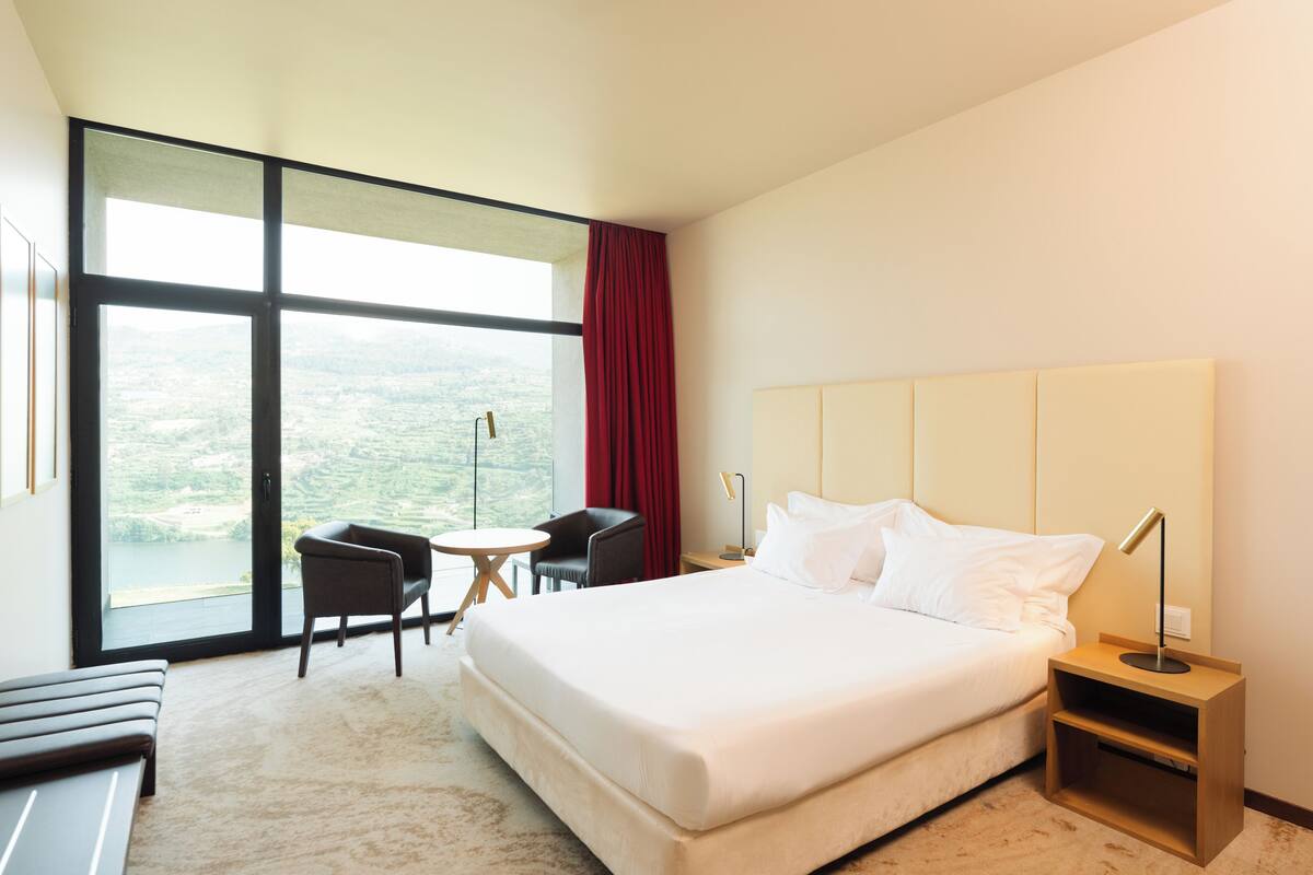 A hotel room with a large bed, a desk, a chair, a small table, and a view of the outdoors.