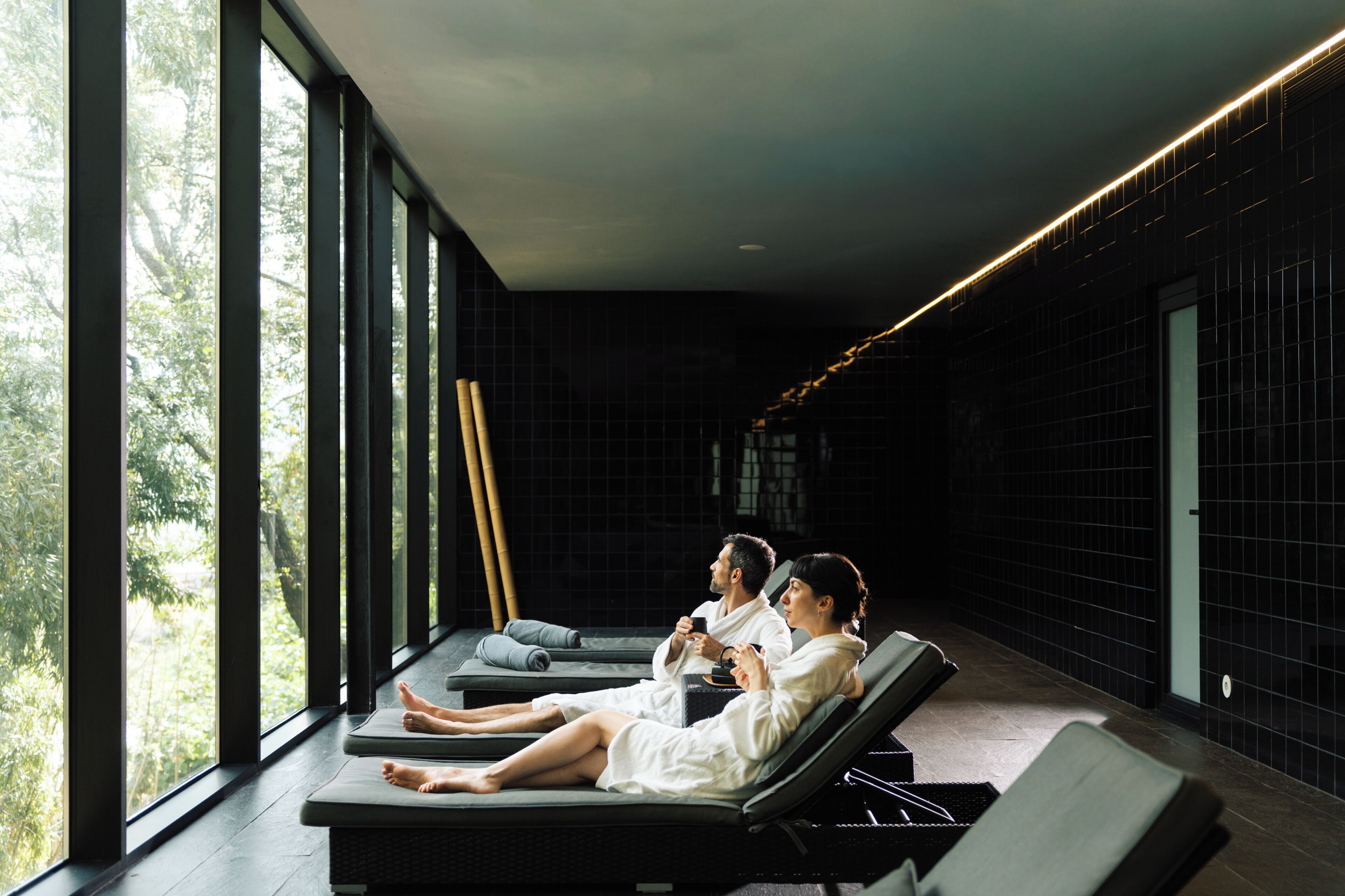 Un cuarto de spa con ventanas amplias, ambiente oscuro y tres personas relajándose en camillas de masaje.