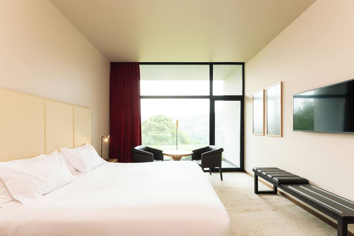 A hotel room with a large bed, a desk, two chairs, a bench, a TV, and a large window.