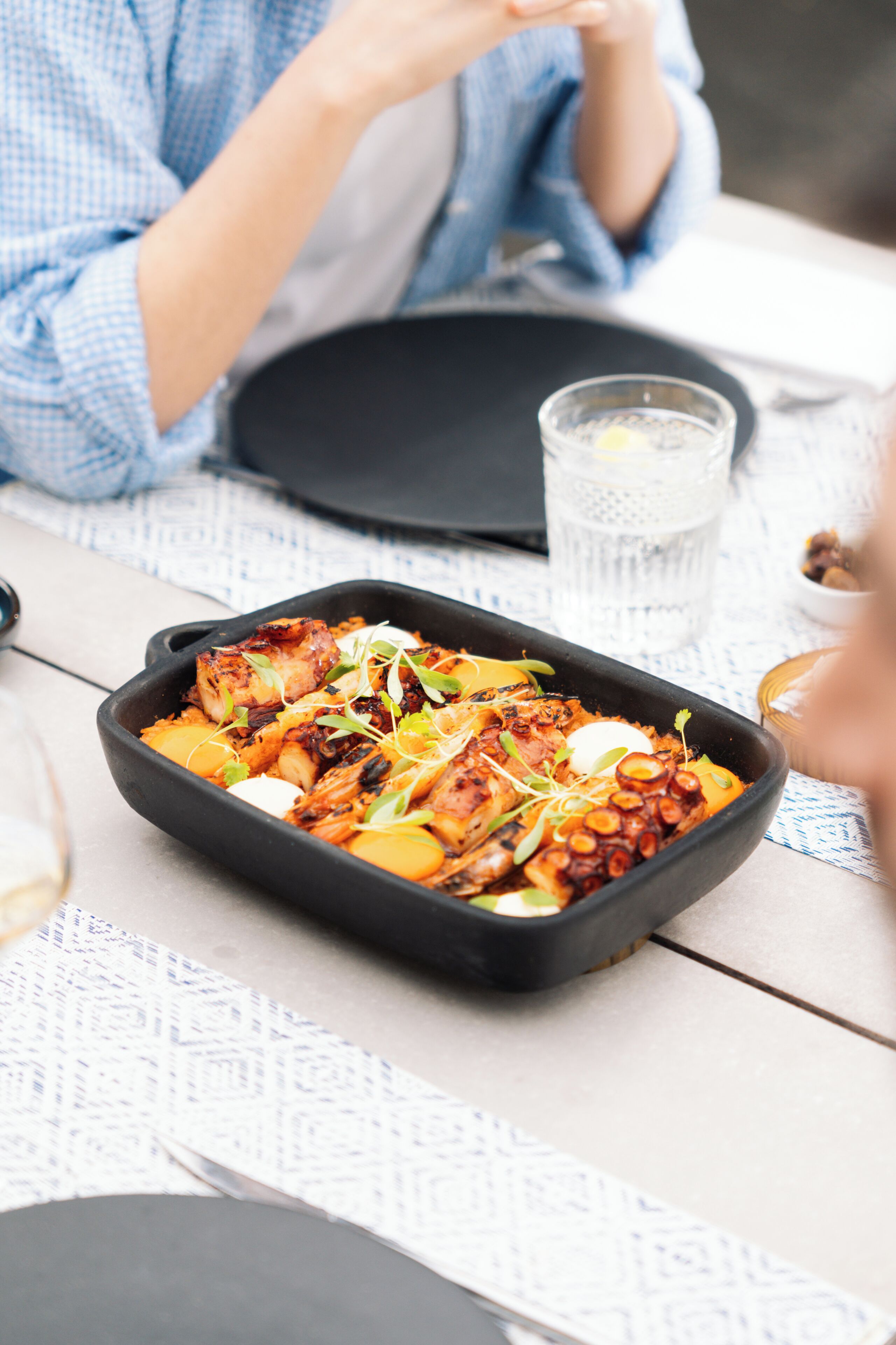 Una mesa de comedor con un recipiente rectangular negro que contiene comida, un vaso de agua y una persona sentada.
