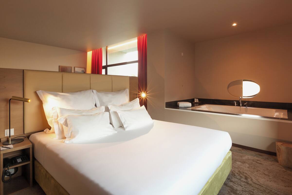 A hotel room with a large bed, a nightstand, a mirror, and a bathroom with a sink and a round mirror.