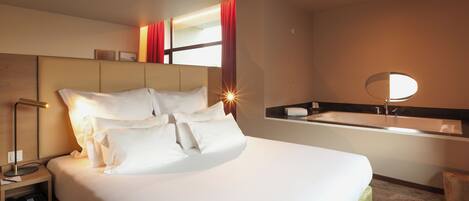 Habitación de hotel con una cama grande, una mesita de noche, un espejo y un baño con lavamanos y un espejo redondo.