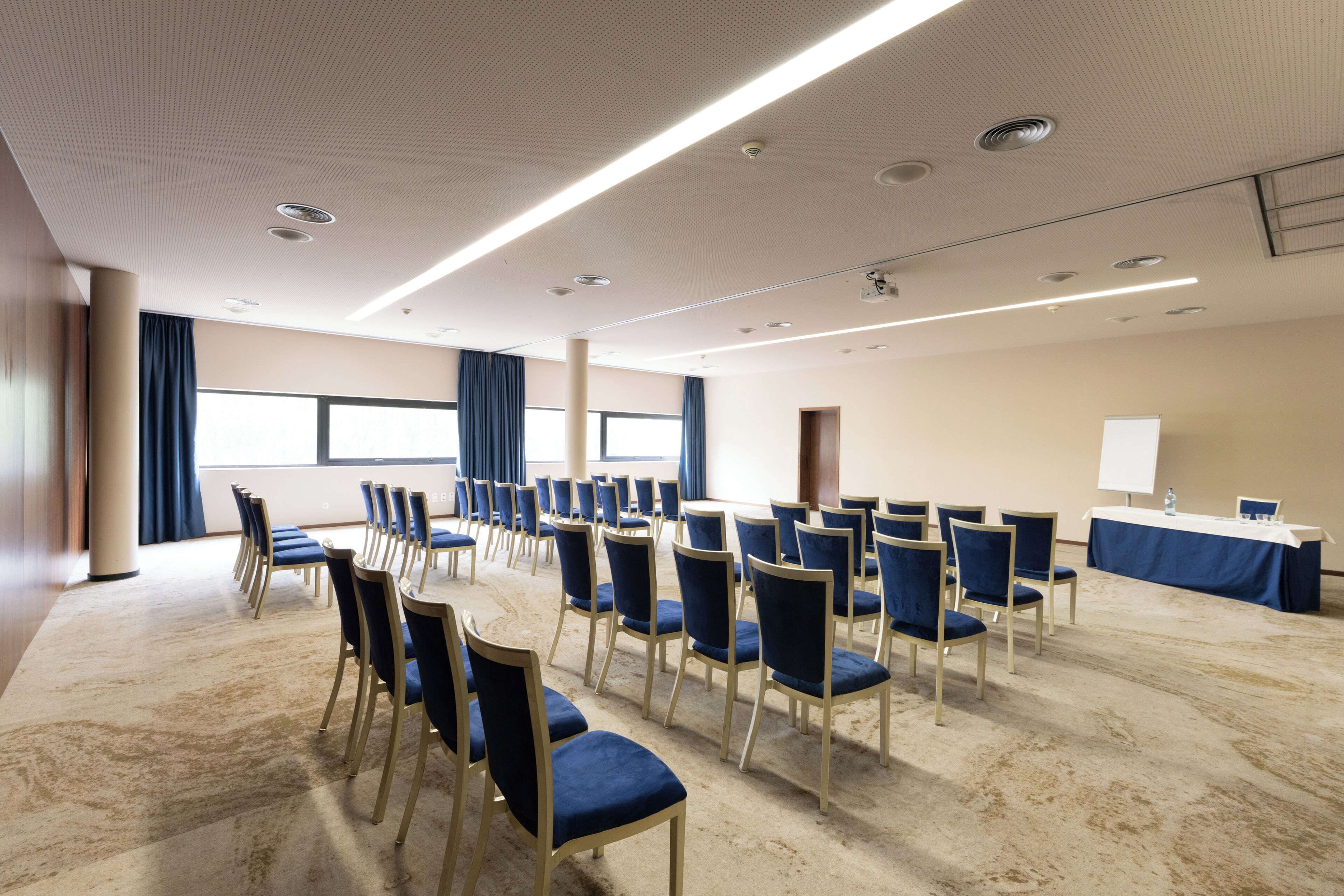 Un salón de conferencias con filas de sillas, una mesa con una pizarra y ventanas grandes con cortinas.