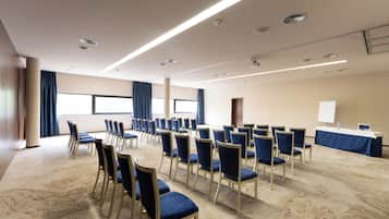 Un salón de conferencias con filas de sillas, una mesa con una pizarra y ventanas grandes con cortinas.
