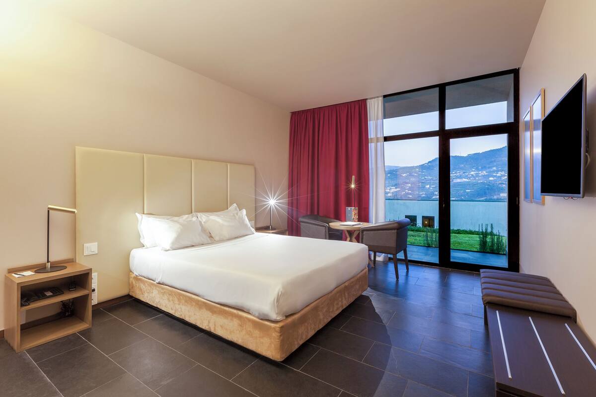A modern hotel room with a large bed, a desk, a chair, a TV, and a view of a body of water.