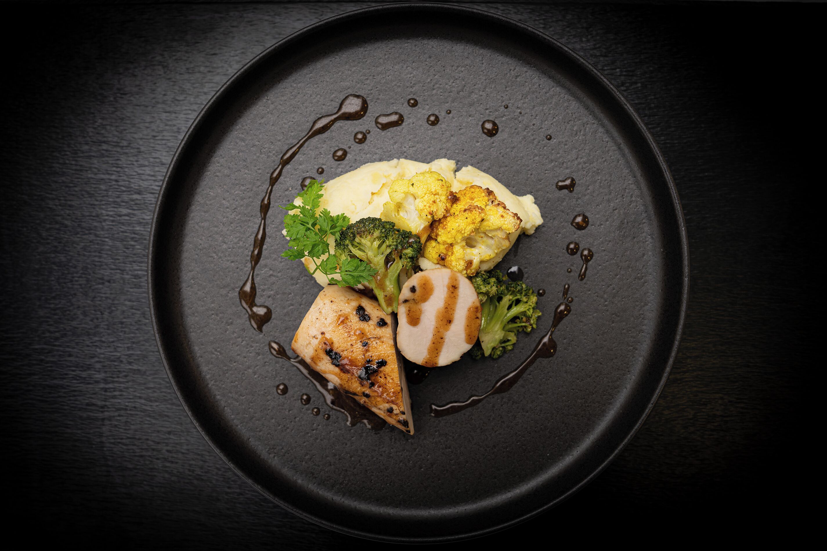 Une assiette noire contenant un morceau de poulet grillé, des brocolis, du chou-fleur et de la purée de pommes de terre.