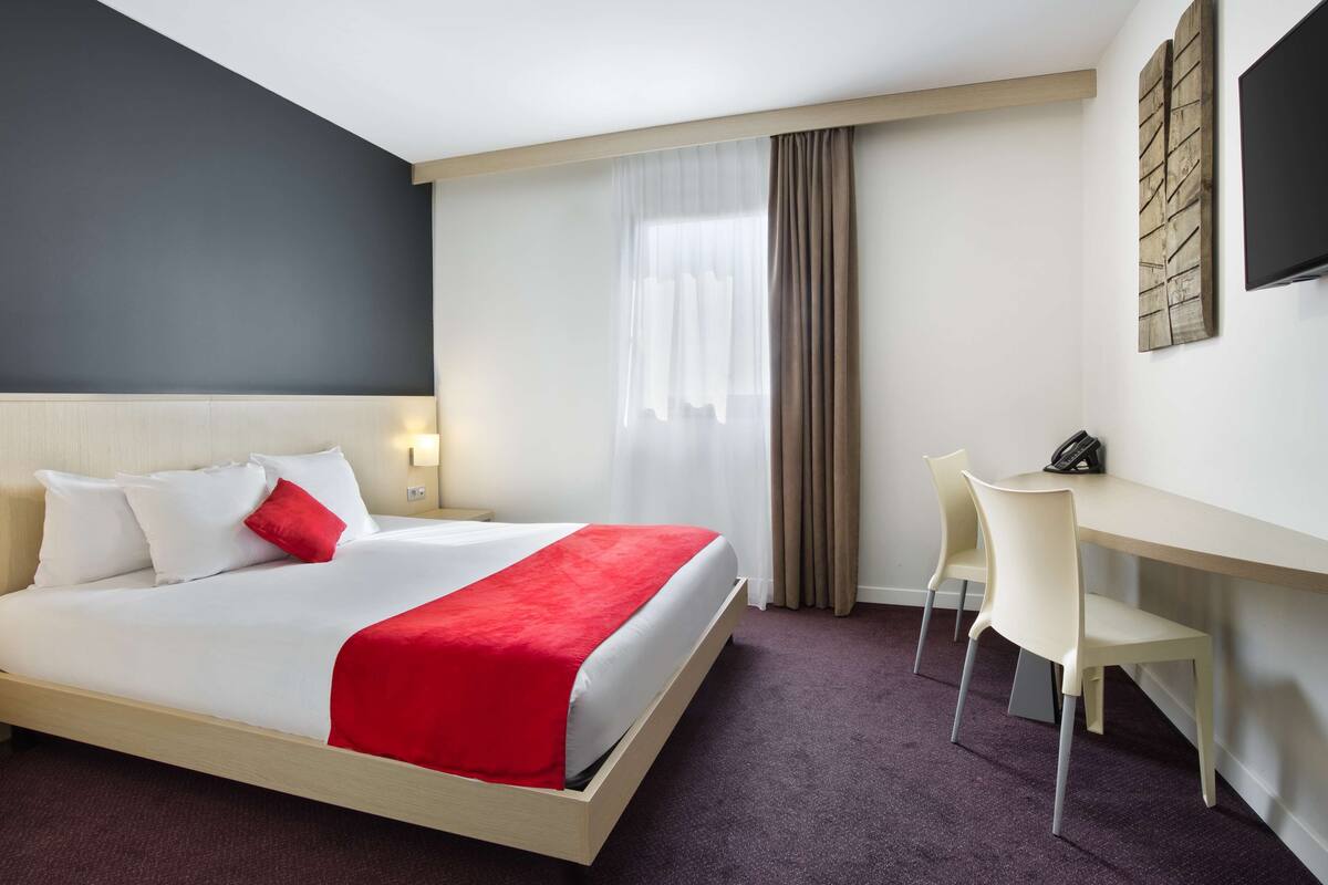 A hotel room with a bed, a desk, a chair, a telephone, and a television.