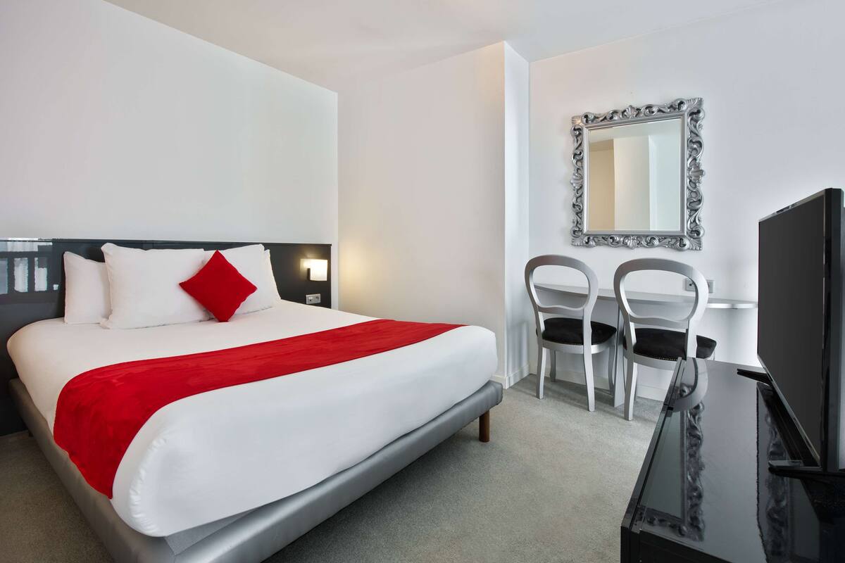 A hotel room with a bed, two chairs, a desk, a television, and a mirror on the wall.