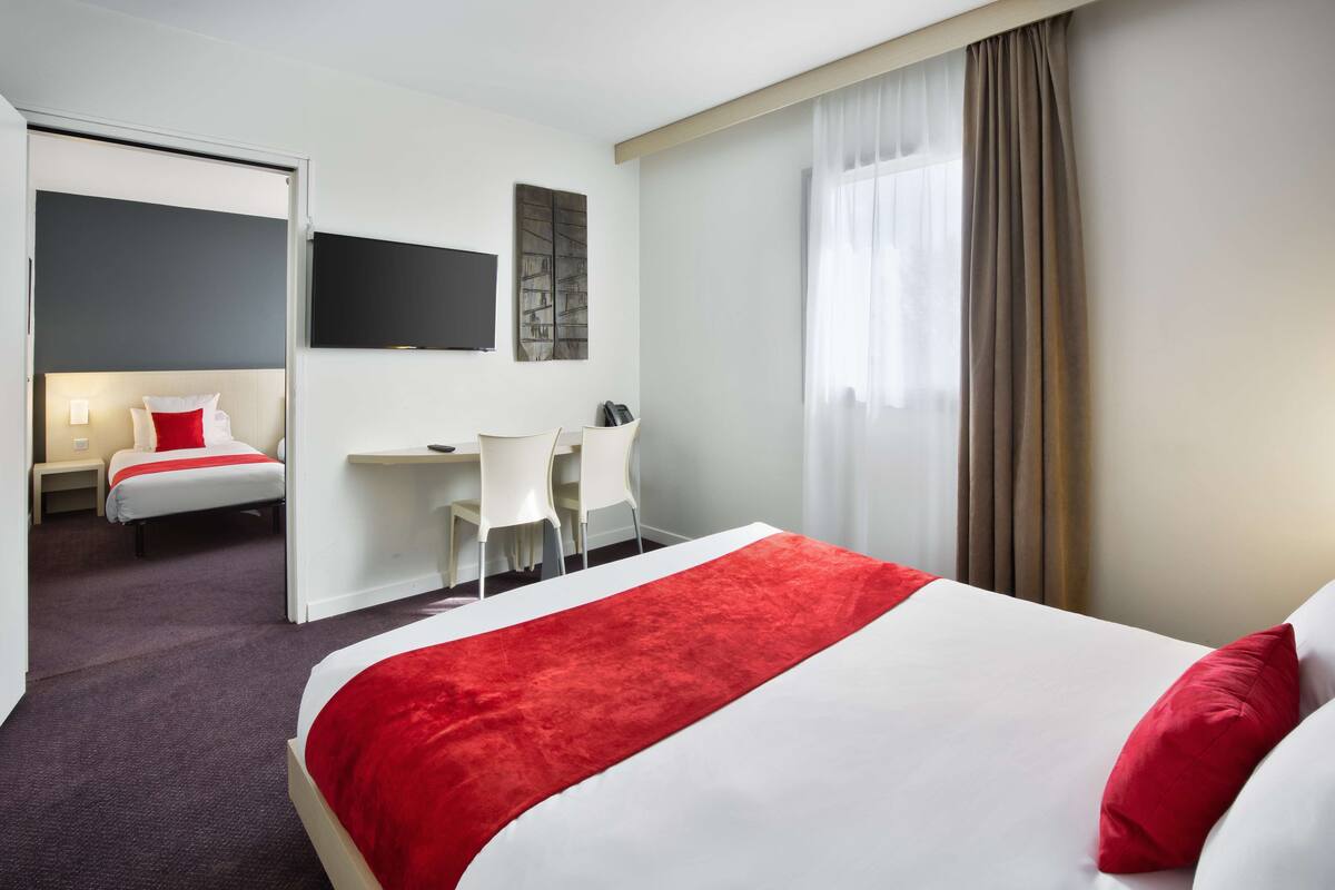 A hotel room with a large bed, a desk, and a chair. There is a TV on the wall and a window with curtains.