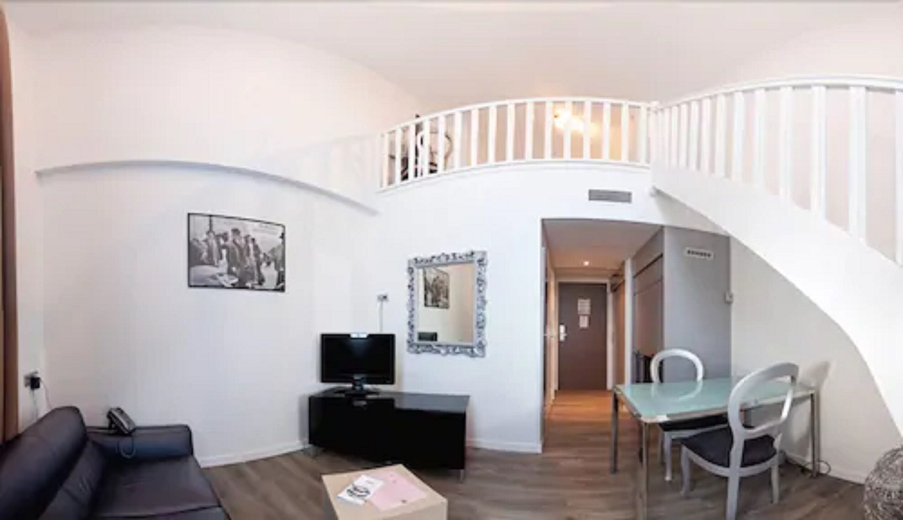 Un salon avec un canapé noir, un téléviseur posé sur un meuble, une table à manger avec des chaises et un escalier menant à l’étage supérieur.