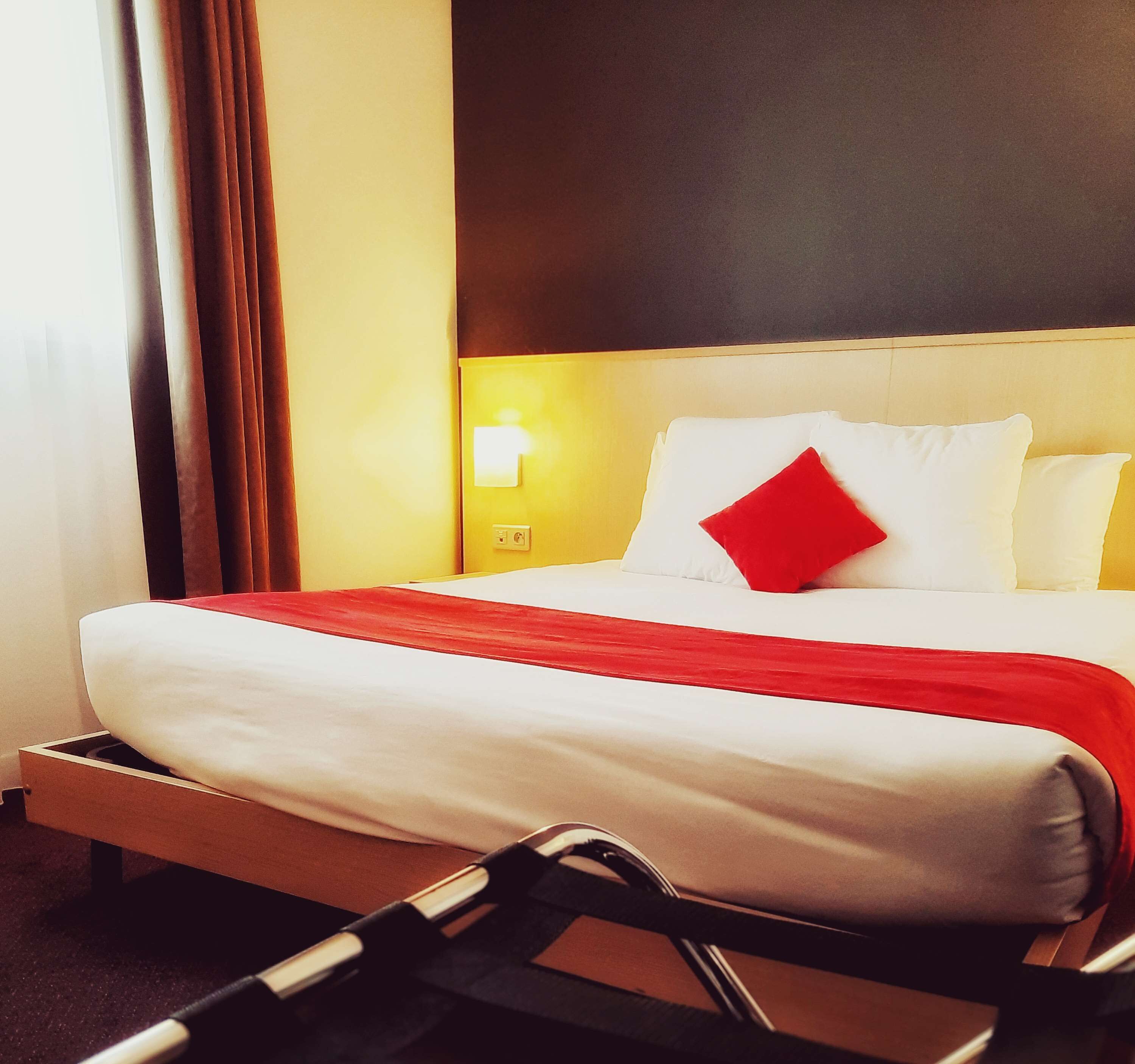 A hotel room with a bed, a red throw, a white pillow, and a red accent pillow.