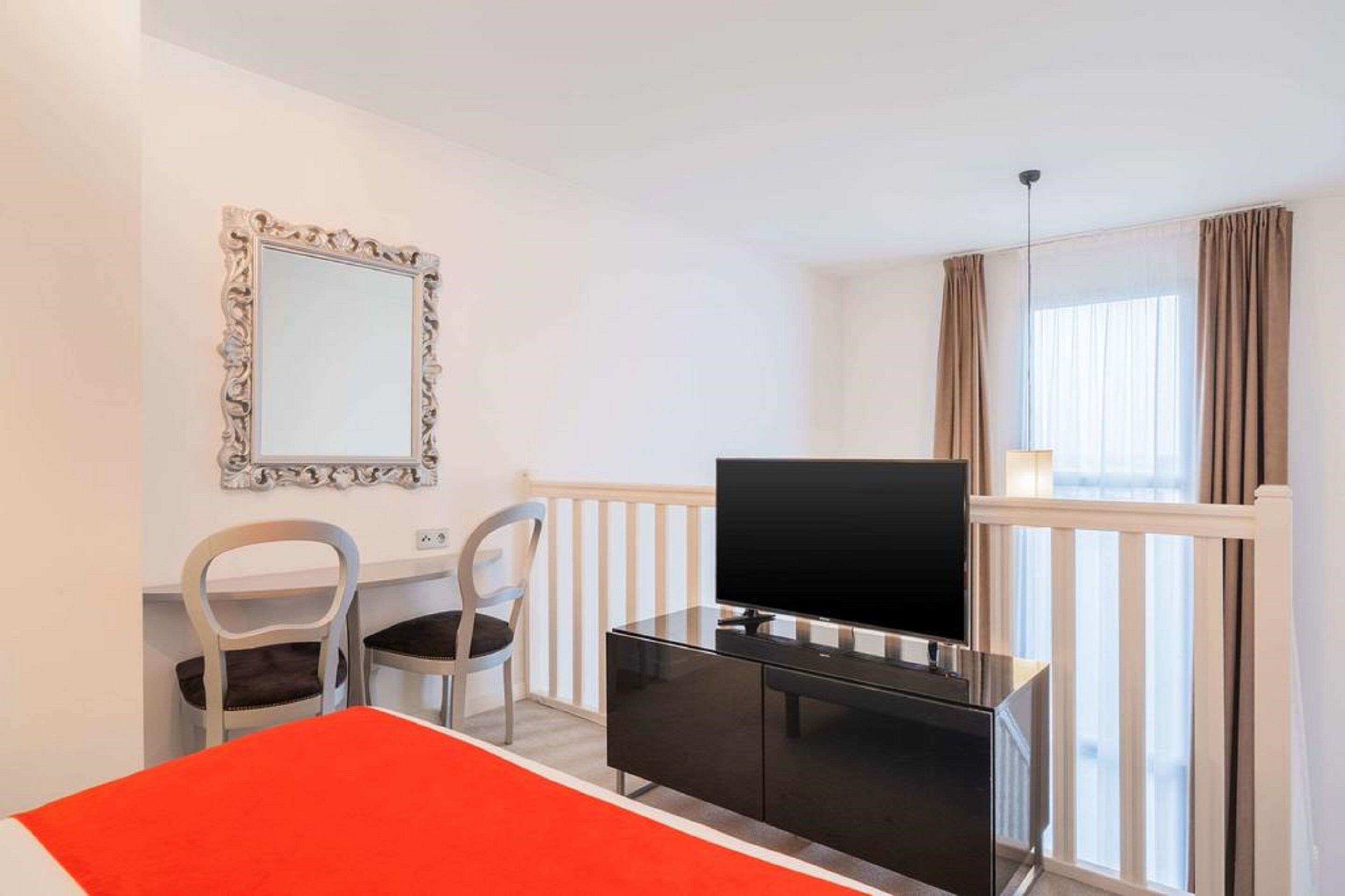 Habitación de hotel con cama, escritorio con televisor, dos sillas, espejo y ventana con cortinas.