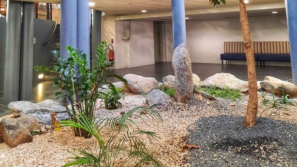 An indoor atrium with a central tree surrounded by rocks and plants, a blue railing, and a seating area in the background. - Sure Hotel by Best Western Nantes Beaujoire (Nantes)