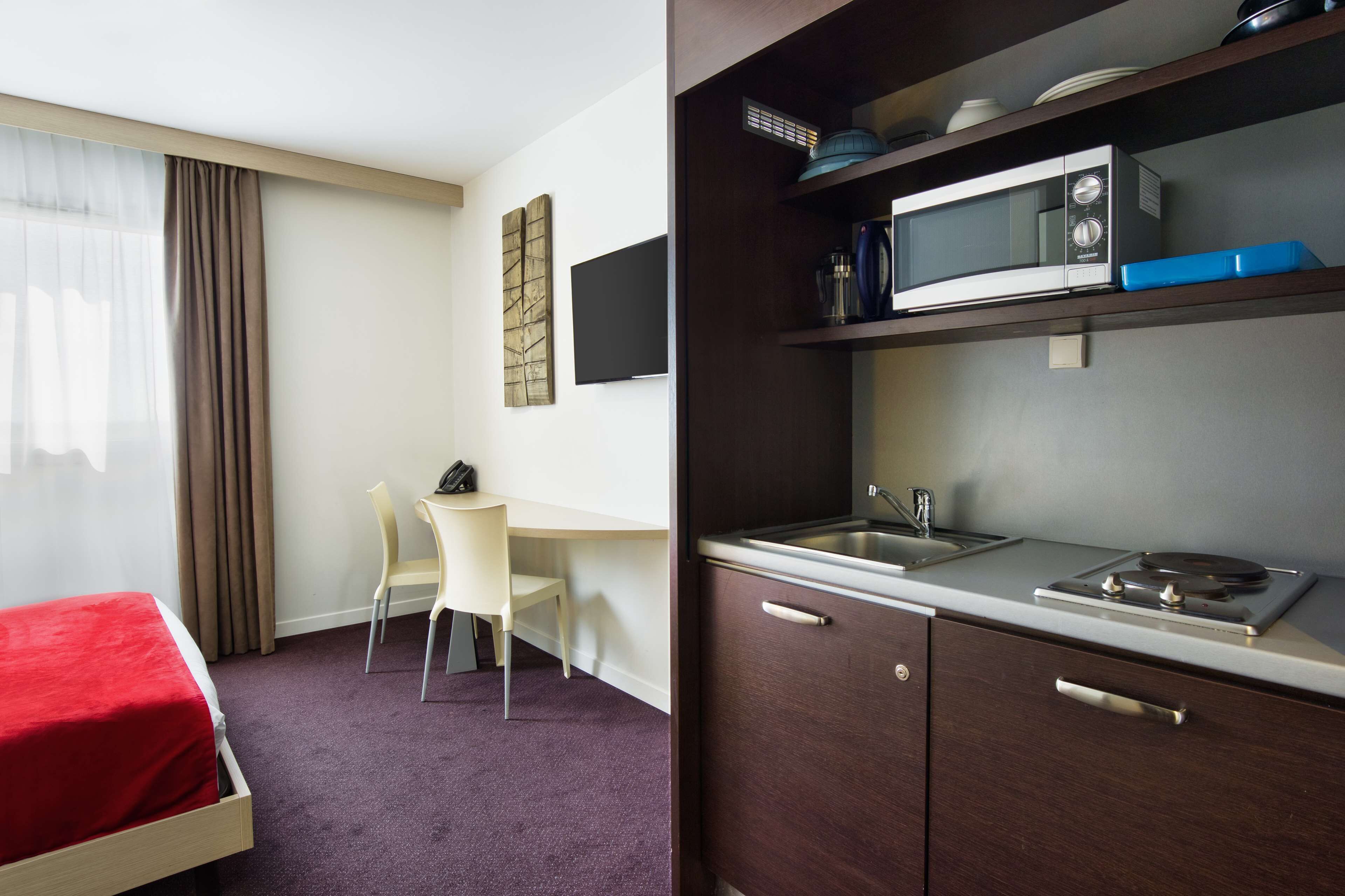 A hotel room with a bed, a desk, a chair, a kitchenette with a microwave, and a sink.