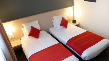 Deux lits simples avec du linge de lit blanc et des touches de rouge, une table de chevet avec une lampe et un luminaire fixé au mur.
