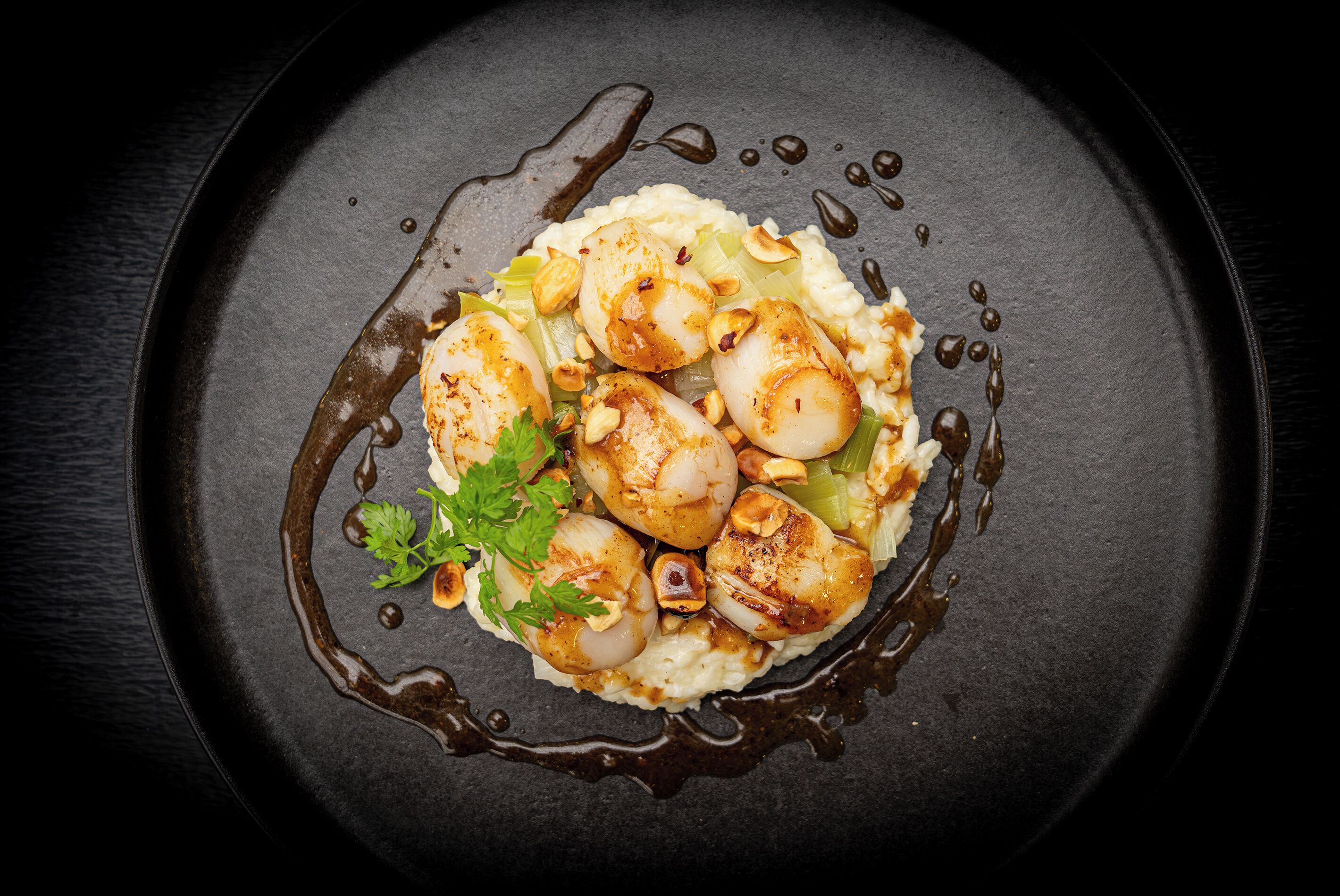 Un plato de comida con vieiras, nueces y perejil sobre un plato oscuro.