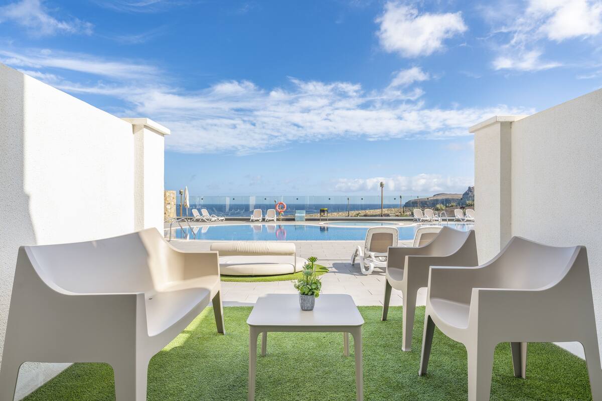 A poolside area with white chairs, a small table, and a view of a swimming pool and distant landscape.