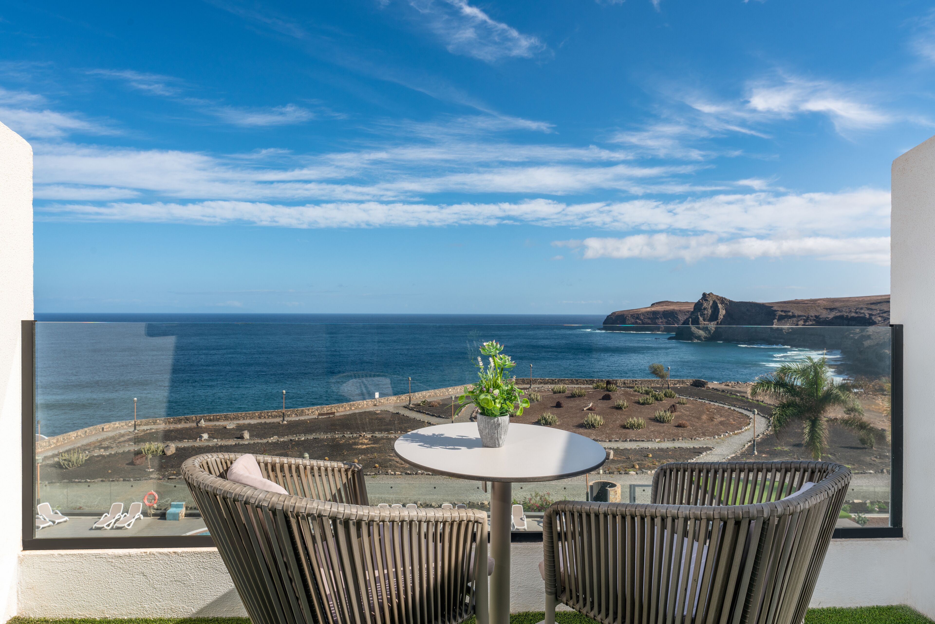 A balcony with wicker chairs, a small table with a plant, and a view of the ocean and rocky coastline.