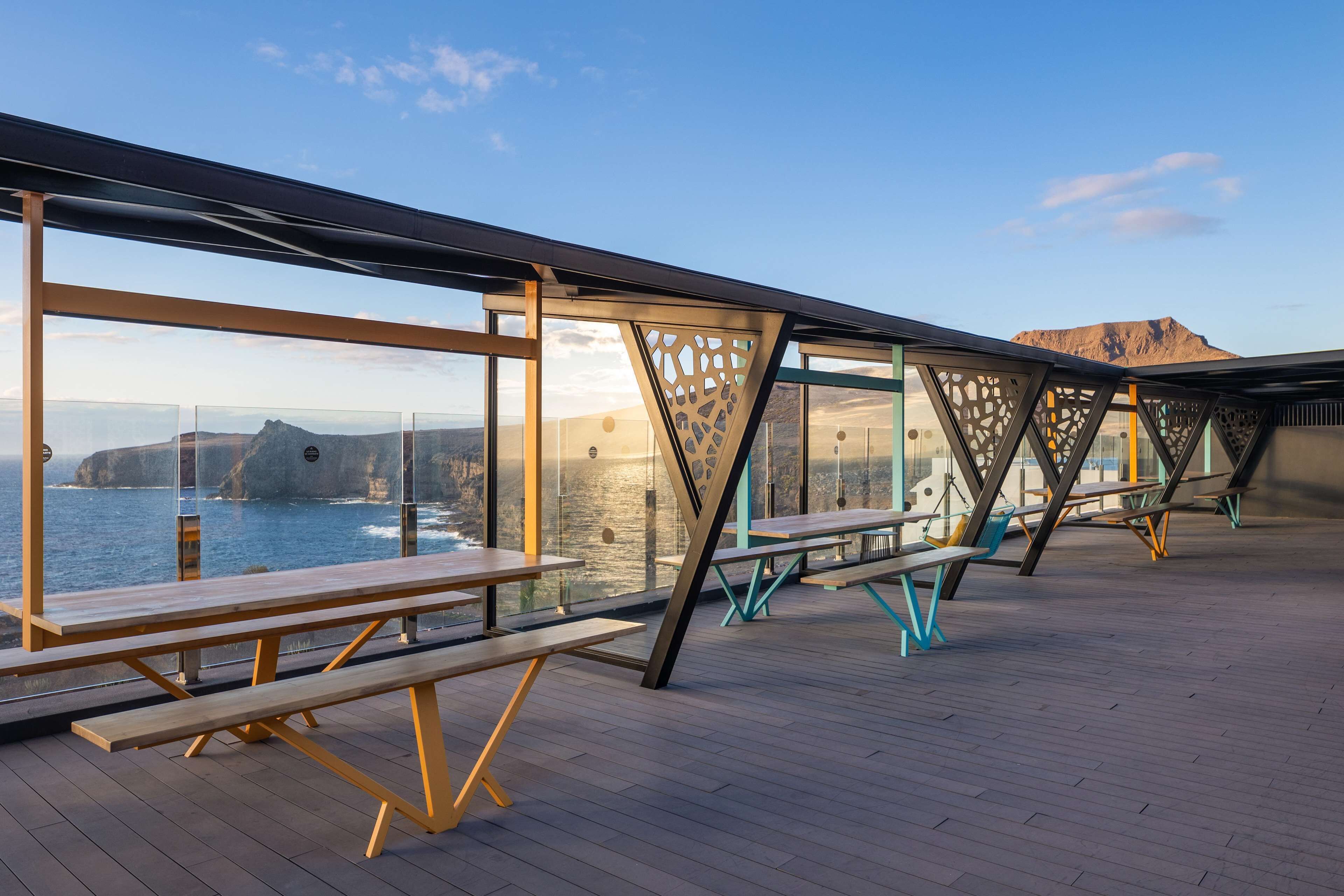 Un espacio al aire libre con bancos y mesas de madera, con vista a un cuerpo de agua y una montaña distante.