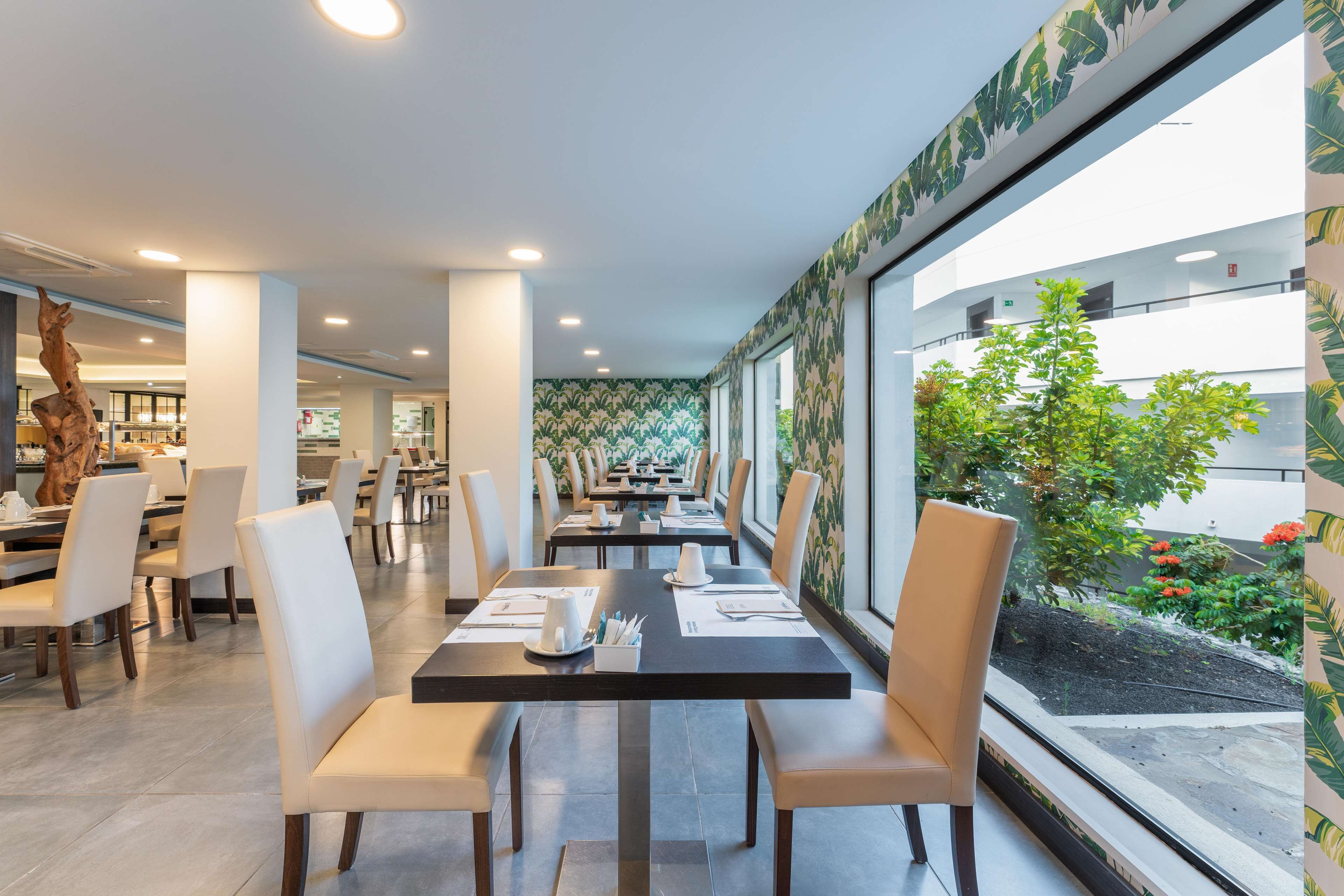 Un restaurant avec de longues tables, des chaises blanches et une vue sur un jardin, grâce à de grandes fenêtres.