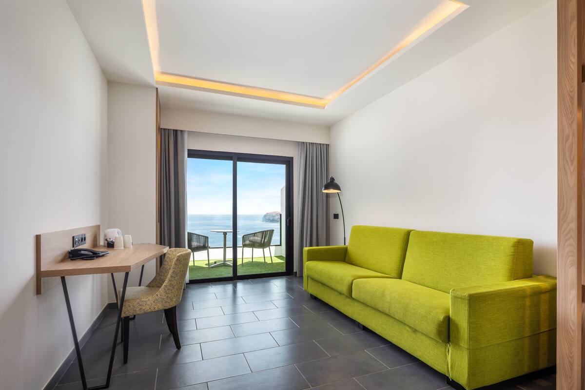 A modern hotel room with a green sofa, a wooden desk, a chair, and a view of the sea through large glass doors.
