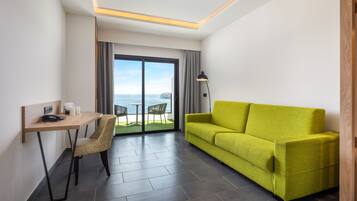 Habitación de hotel moderna con sofá verde, escritorio de madera, una silla y vistas al mar a través de amplios ventanales.