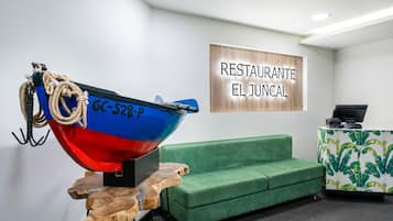 Un espace d’accueil avec une enseigne en bois portant l’inscription « RESTAURANTE EL JUNCAL », une sculpture représentant un bateau aux couleurs bleu et rouge, un canapé vert et un meuble orné de motifs.