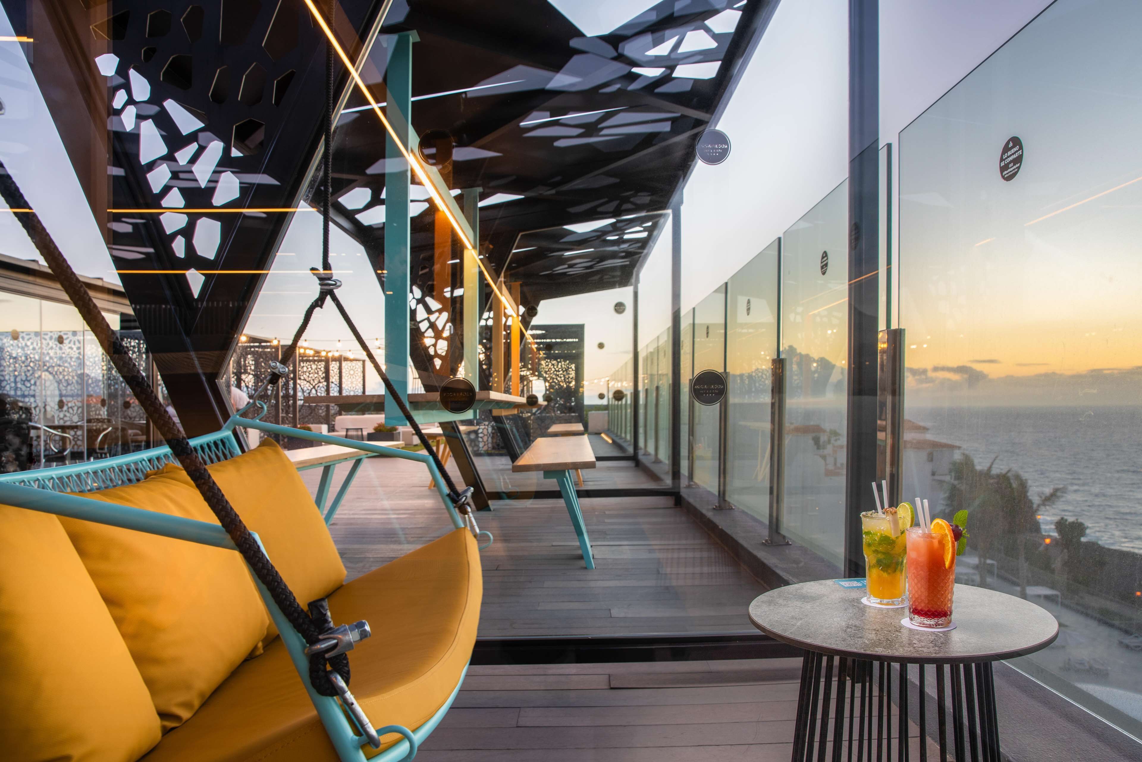 Un espace extérieur moderne aménagé pour se détendre, avec vue sur l’océan, comprenant un fauteuil suspendu, une table avec des boissons et une rambarde en verre.