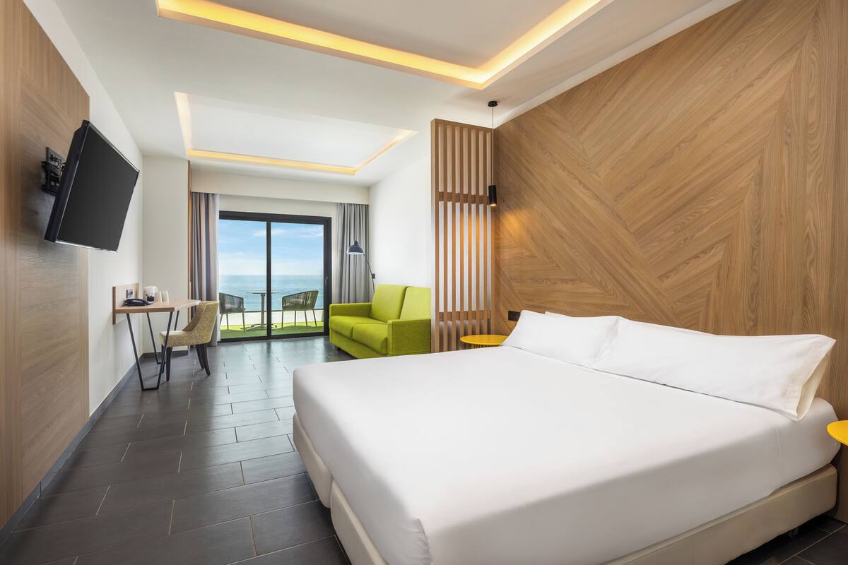 A modern hotel room with a bed, a desk, a chair, a TV, and a view of the outdoors.