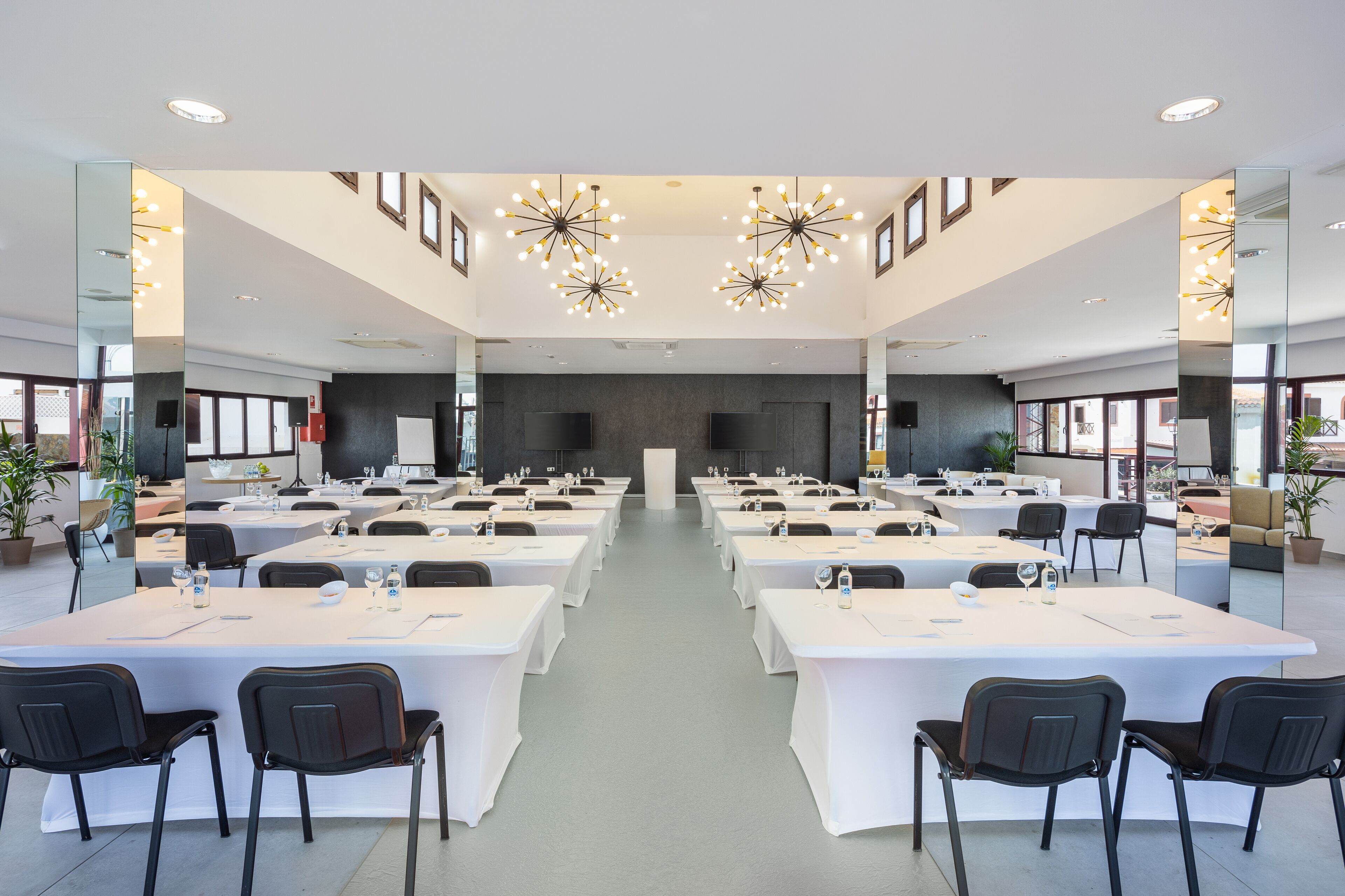 Un amplio salón de conferencias con varias mesas y sillas, candelabros modernos y ventanas grandes.