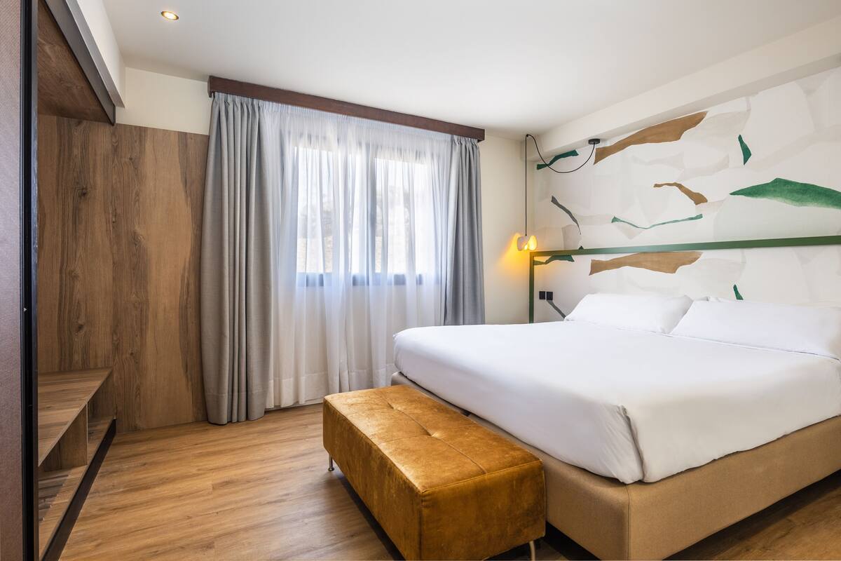 A modern hotel room with a wooden floor, a large bed with white bedding, a bench, a window with curtains, and a wall with abstract art.