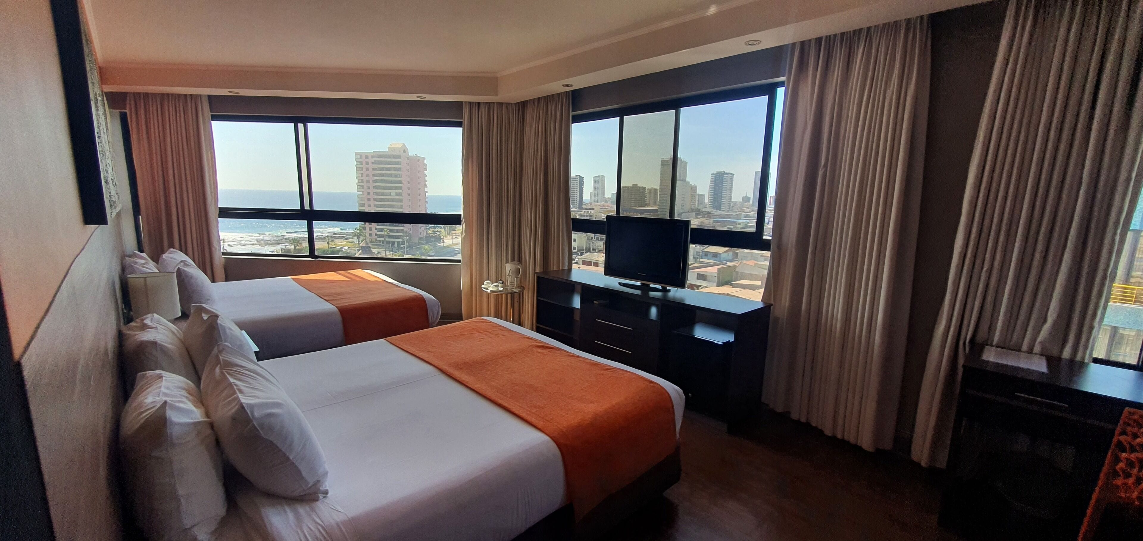 A hotel room with two beds, a TV, a desk, and large windows offering a city view.
