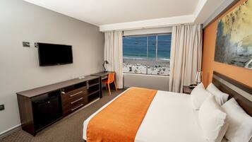 A hotel room with a bed, a desk, a TV, and a view of the beach.
