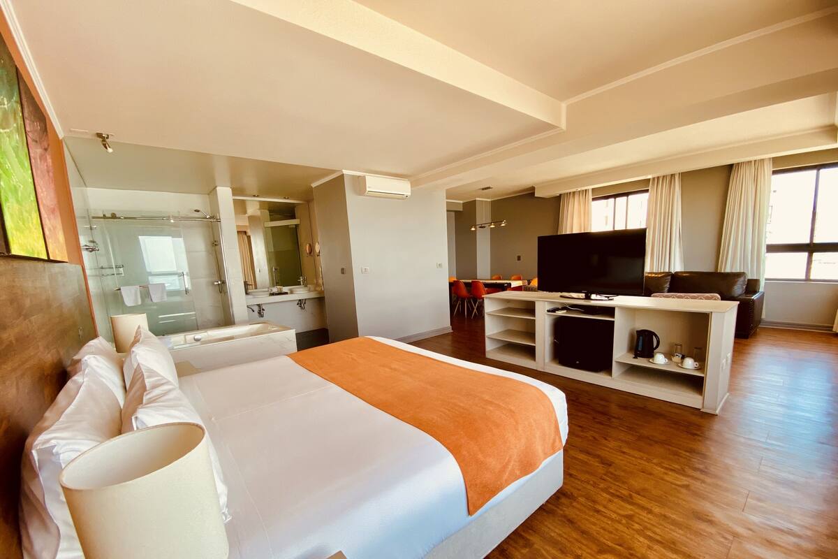 A hotel room with a large bed, a TV, a seating area, and a bathroom with a mirror and sink.