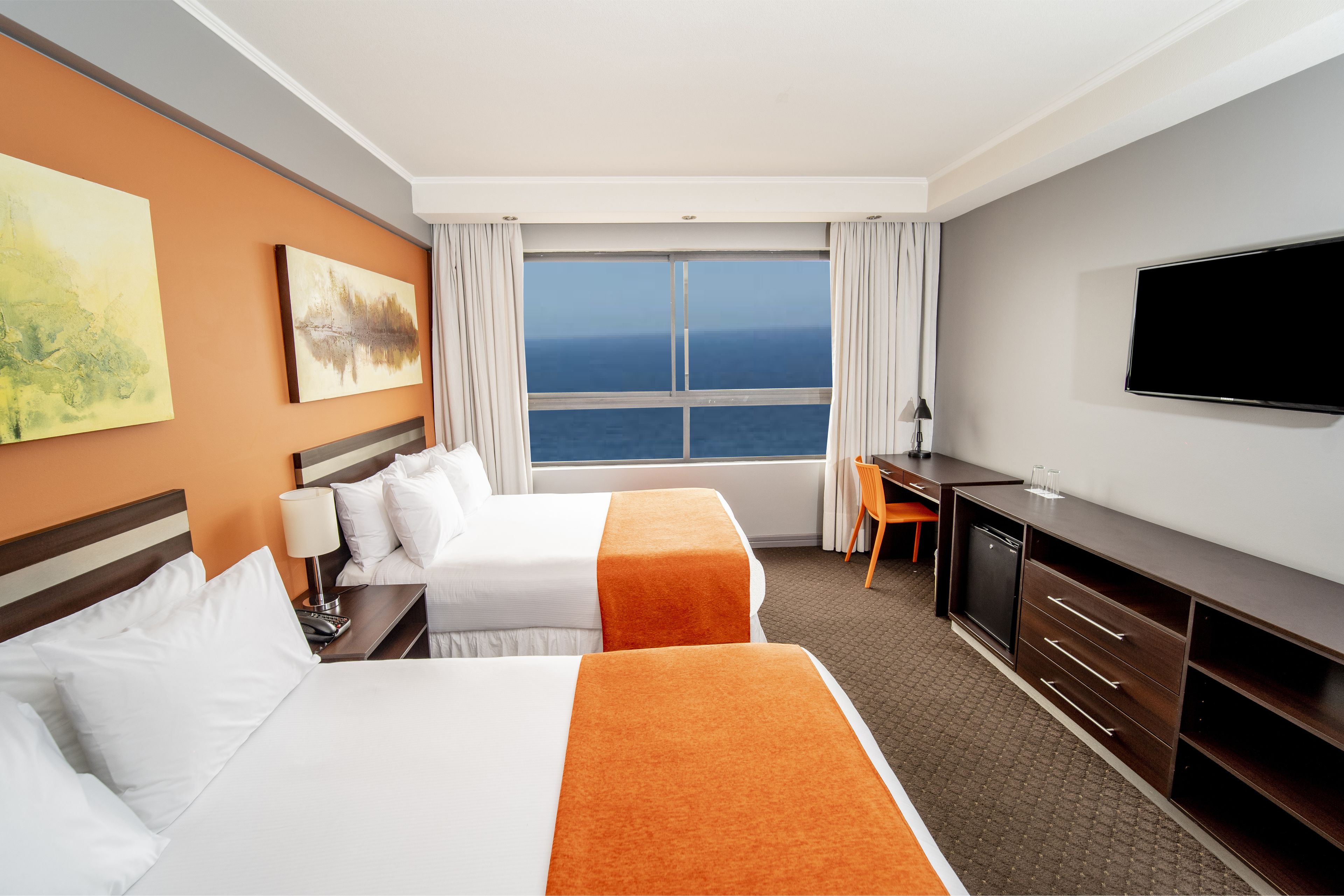 A hotel room with two beds, a desk, a TV, and a large window with a view of the sea.